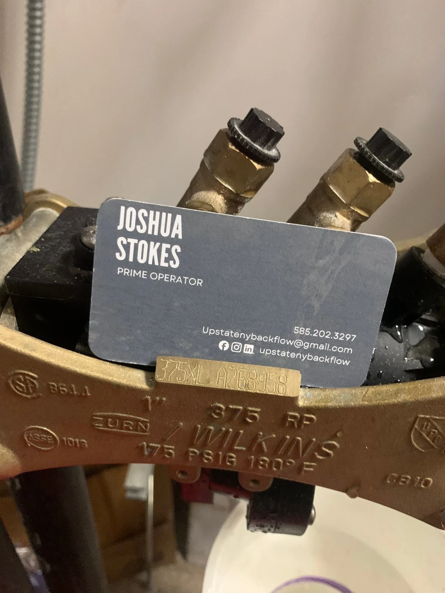 Upstate NY Backflow business card for Joshua Stokes resting on a Zurn Wilkins 375 reduced pressure assembly